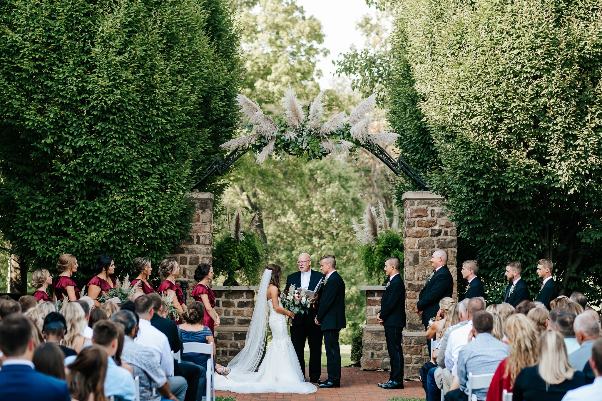 Bride and Groom at alter in a garden with lush greenery and trees. Many guest watching event.