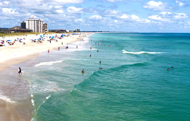 Wrightsville Beach Coastline with blue green white sandy shore coast with beach goers sunbathing and in water.