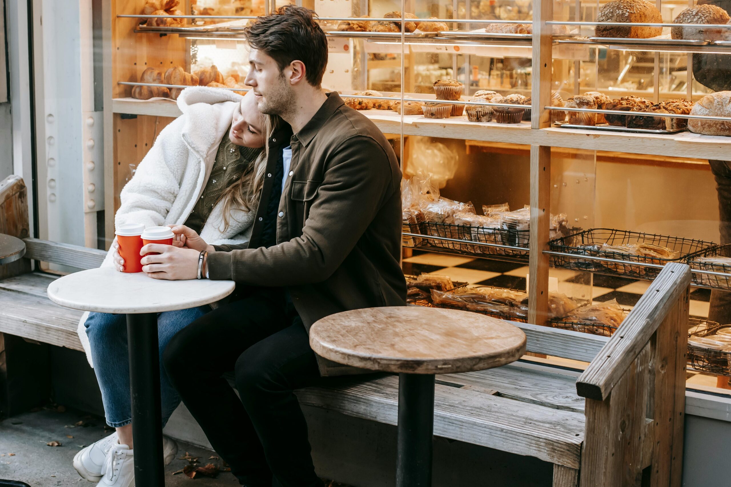 Photo of a man drinking coffee in a coffee shop with baked goods in a glass case behind him.