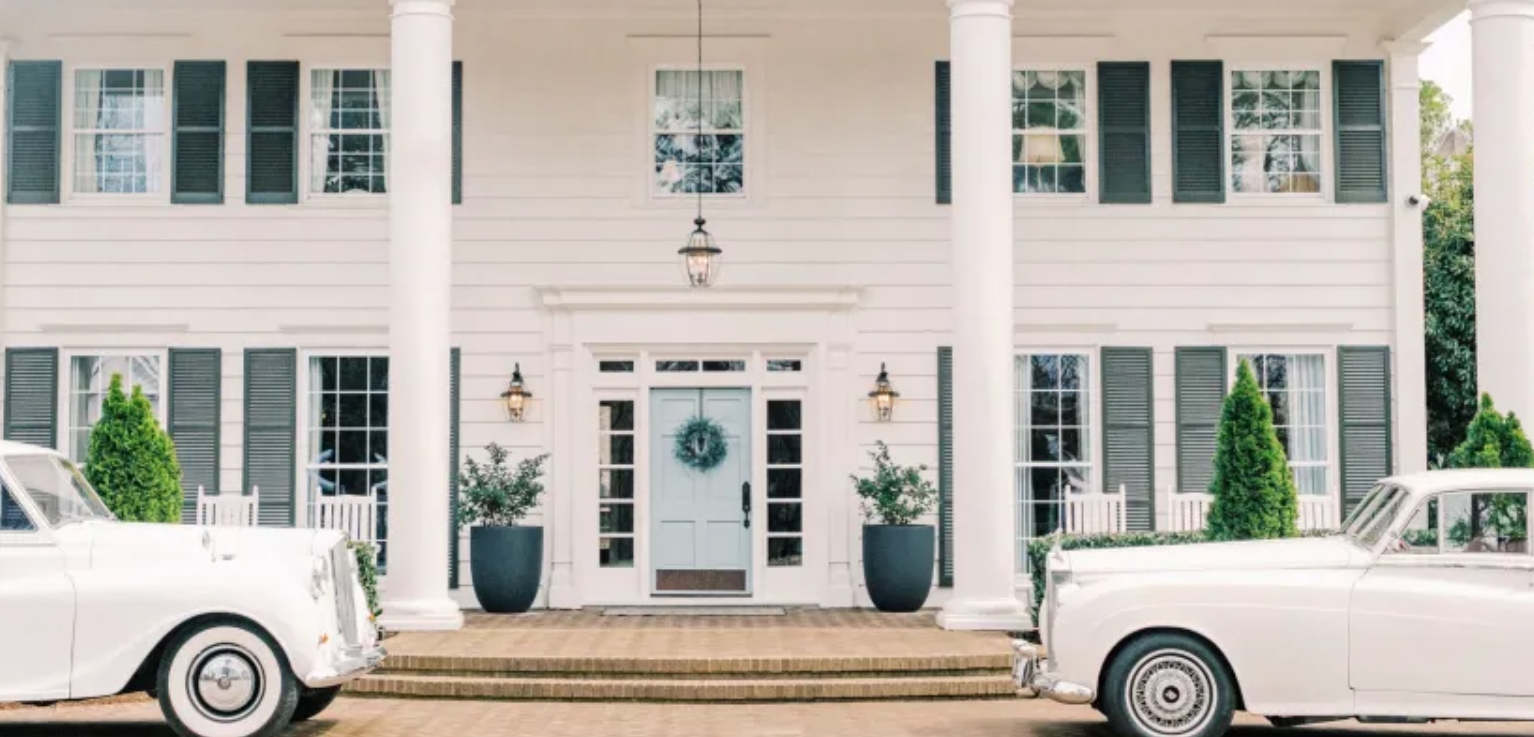 Highgrove Estate Entrance white painted exterior with large wood columns flanking the door. Shutters on the exterior of the windows. 2 Rolls Royce Cars parked in front of a stone drive.