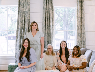 Photo of a group of ladies that own a wedding venue in Wilmington NC. Posing on a large gray sofa and printed floral draperies flanking the windows. Shiplap as the wallcovering.