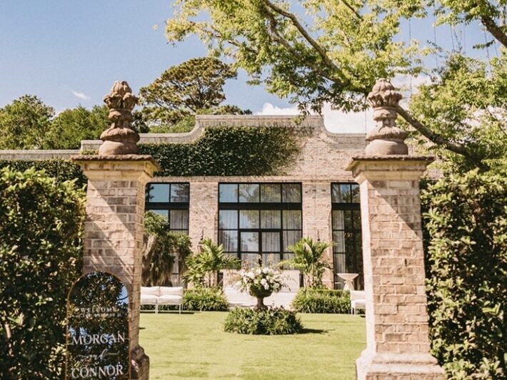 Facade of the venue with large stone columns flanking the garden area. Large finials on top of columns. Lush green grass and Ivy adorning the stone building. Lawn furniture adorning the large courtyard.