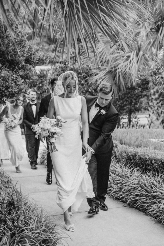 Couple and wedding part walking out of the ceremony space at Wrightsville Manor after getting married. Groom helping bride with dress.