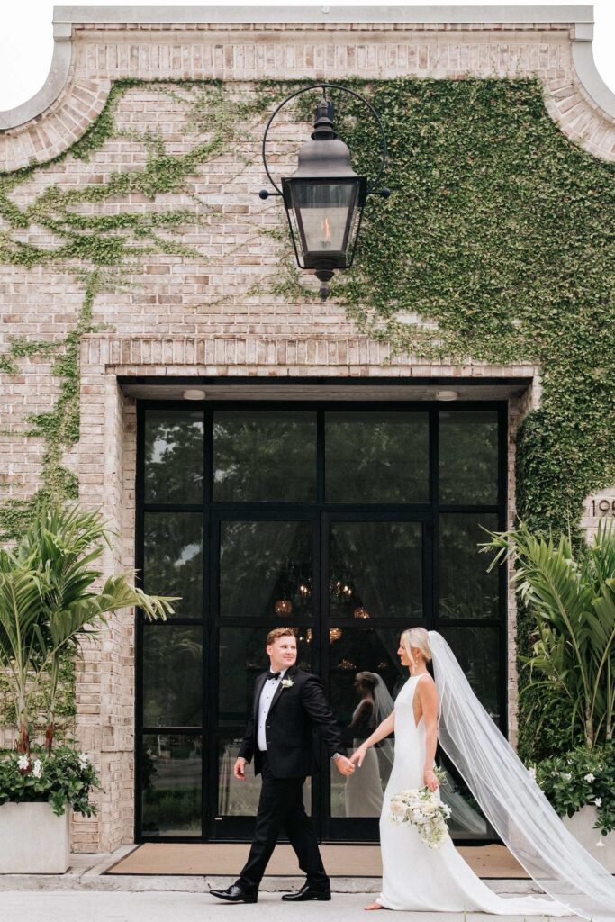Couple walking and holding hands in front of the window with brick and greenery in the background at Wrightsville Manor wedding.